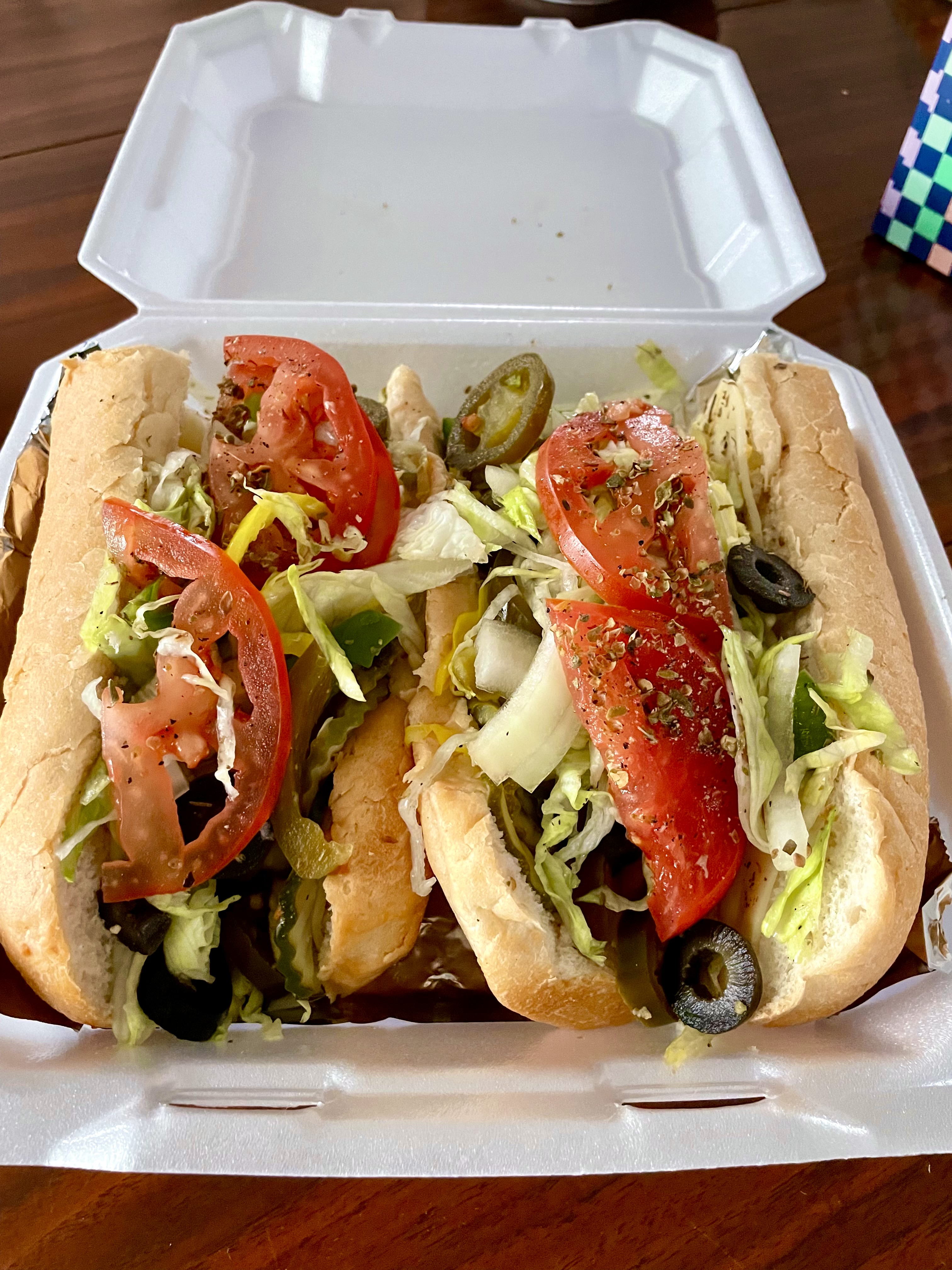A close-up of a sub topped with tomatoes, lettuce and olives in a takeout container